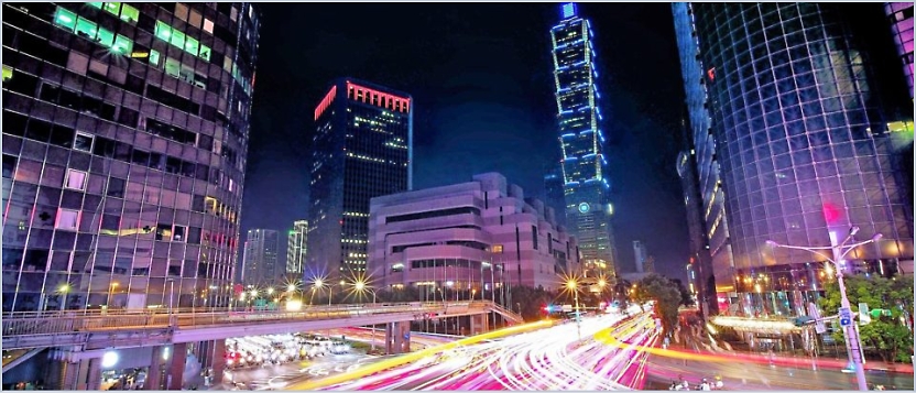 Nighttime cityscape with tall, illuminated buildings and vibrant light trails from moving vehicles on the streets below.