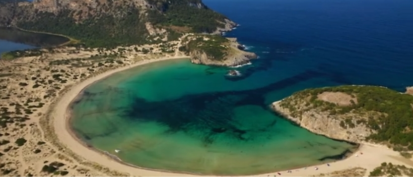 Flygfoto över en halvmåneformad strand med klart turkos vatten, omgivet av klippiga och gröna landskap.