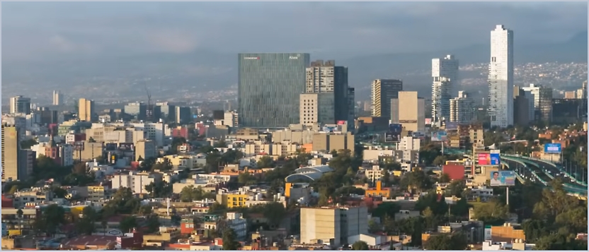 Aerial view of a cityscape featuring numerous buildings and skyscrapers under a cloudy sky, with mountains in the distant background.