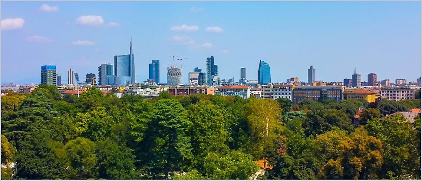 Panoramic view of a city skyline in the background and abundant green trees in the foreground under a clear blue sky.