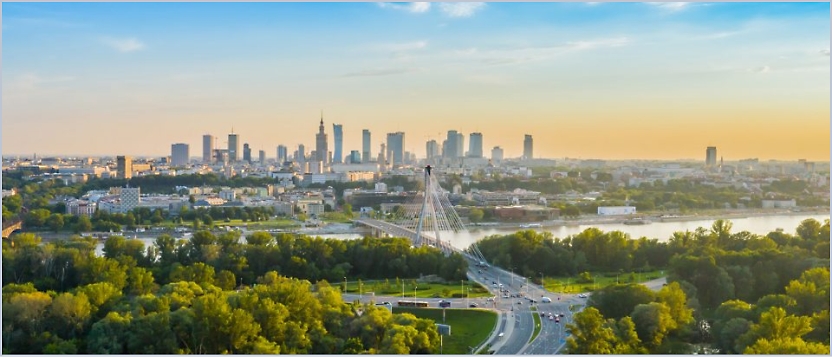 Aerial view of a city skyline with tall buildings in the background, a river, and a bridge surrounded by green trees in the foreground