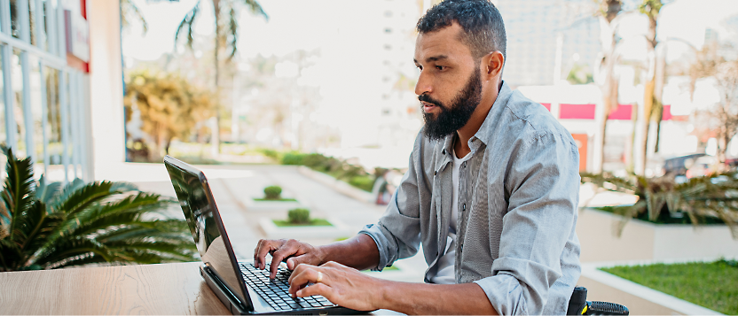 Man with a beard and mustache sitting outdoors, working on a laptop, with buildings and greenery in the background.