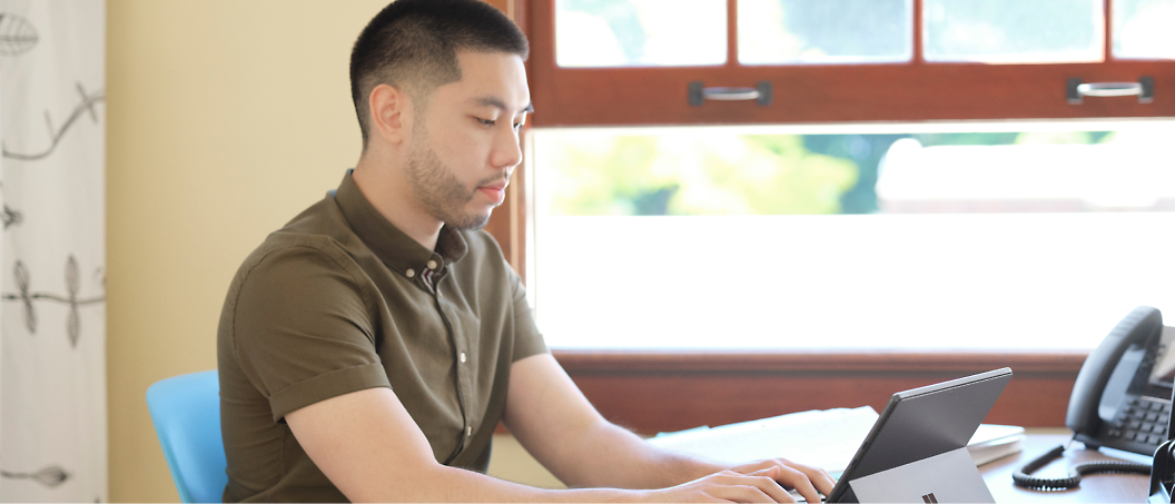 A man is seated at a table, working on a tablet in a bright room with a window.