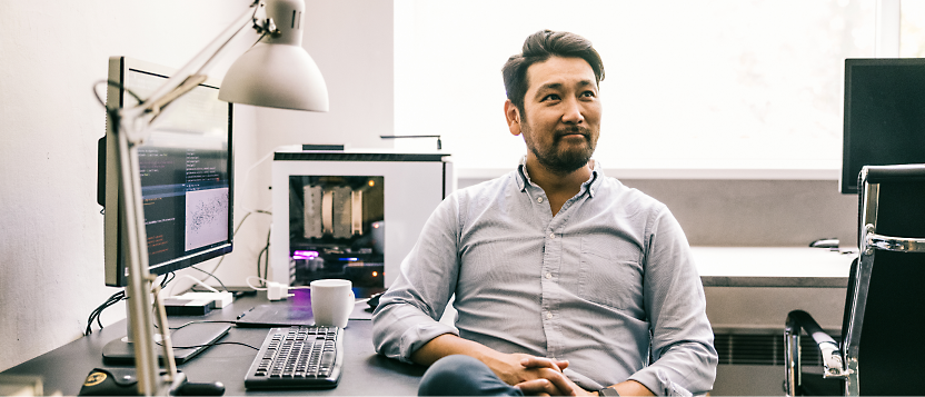 Asiatisk man i en blå tröja som sitter vid ett skrivbord med datorer och tittar eftertänksamt åt sidan på ett väl upplyst kontor.