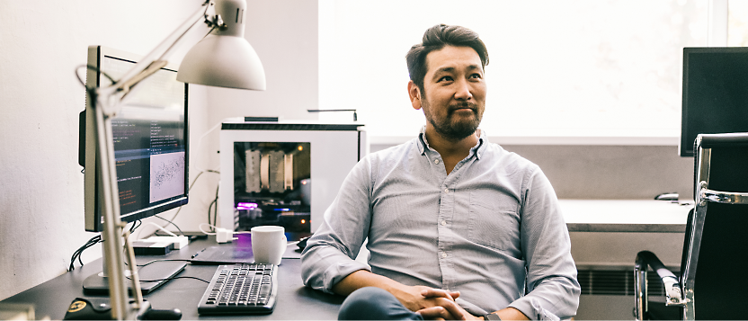 Asian man in a blue shirt sitting at a desk with computers, looking thoughtfully to the side in a well-lit office.