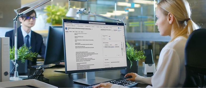 Two people in an office setting: foreground individual working on a desktop computer displaying a detailed interface.