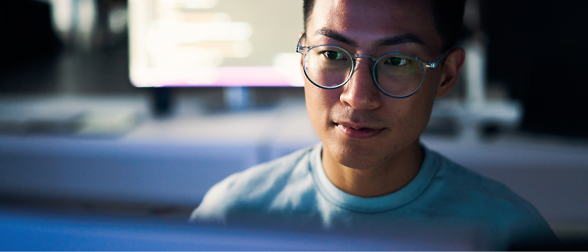 Ung asiatisk man med glasögon som arbetar koncentrerad på en dator i ett dåligt upplyst rum, med fokus på ansiktet