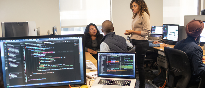 A diverse team of five professionals collaborates in an office setting, with one woman standing and presenting to her 