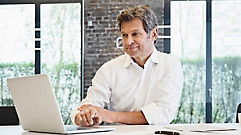 A man sits at a table, working on a laptop, with a brick wall, large windows, and greenery in the background.