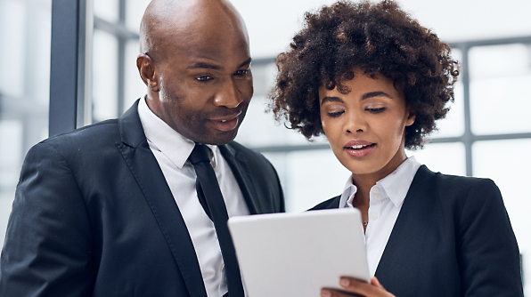 Two professionally dressed individuals are looking at a tablet together in an office setting.