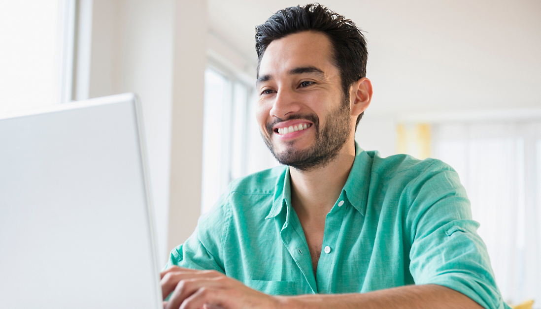 A man in a green shirt is smiling while using a laptop.