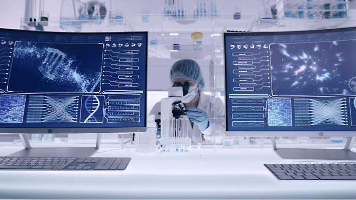 A person looking through a microscope at a desk with screens displaying DNA and more