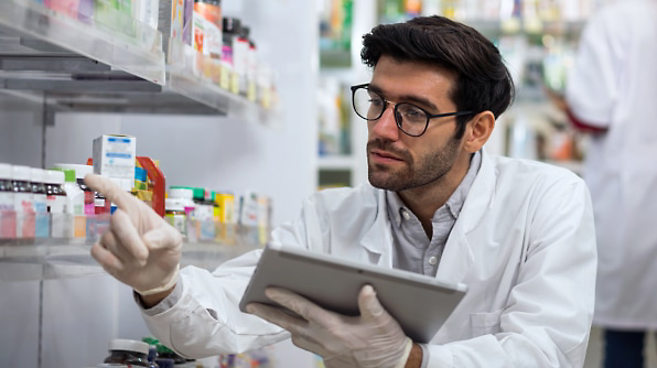 A pharmacist wearing a white coat and gloves holds a tablet while selecting a medication from a shelf in a pharmacy.