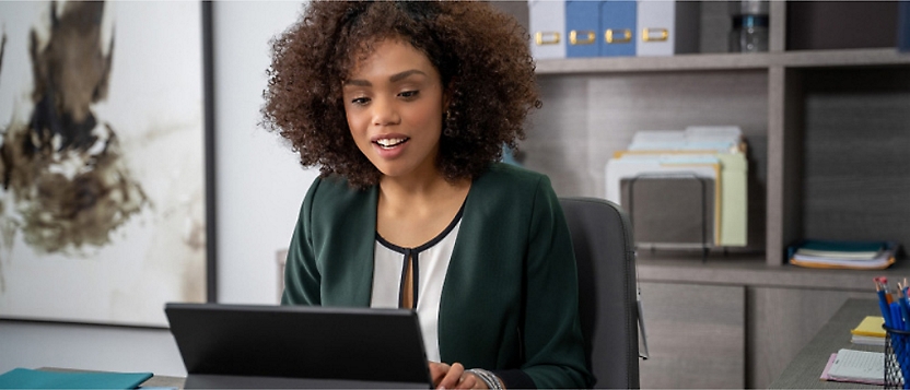 A woman with curly hair wearing a green blazer sits at a desk working on a laptop 