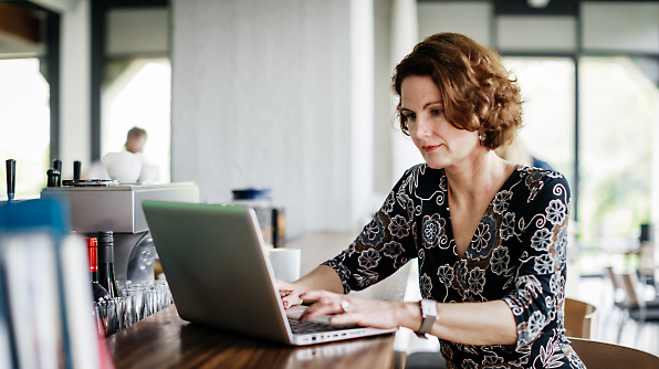 A woman with short curly hair is sitting at a wooden counter in a cafe, typing on a laptop. 