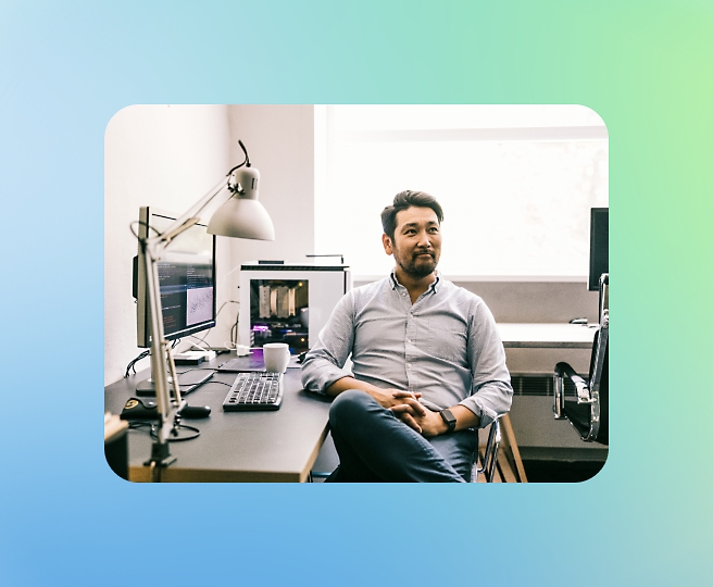 A man in a light blue shirt sitting at a desk with computer monitors, a lamp, and a coffee mug, in a brightly lit room.