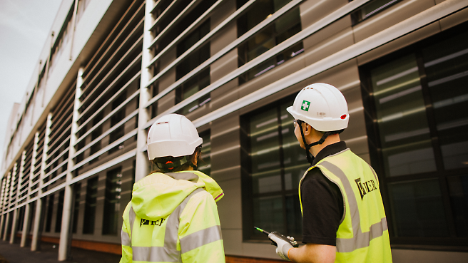 Two construction workers wearing safety vests and helmets are looking towards a modern building.