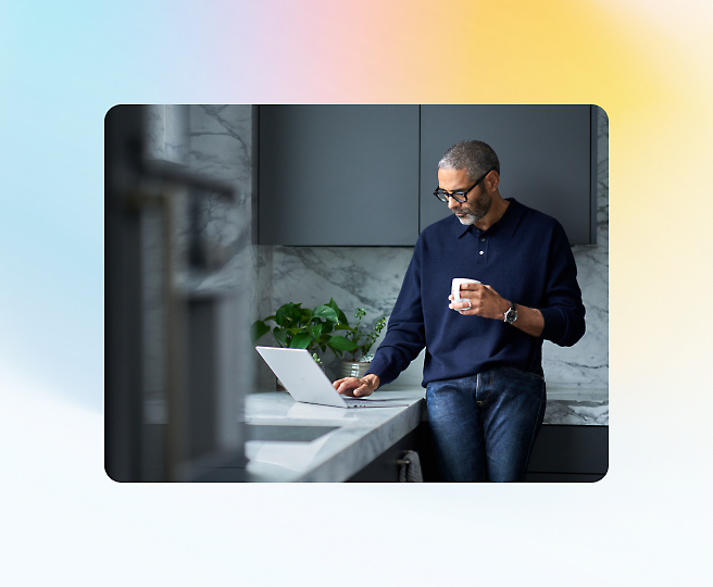 A person in a navy sweater stands in a modern kitchen, holding a mug and looking at a laptop on the counter.