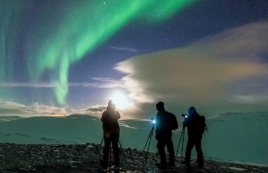 Drei Personen stehen mit Kameras unter grün-blau gefärbten Polarlichtern.