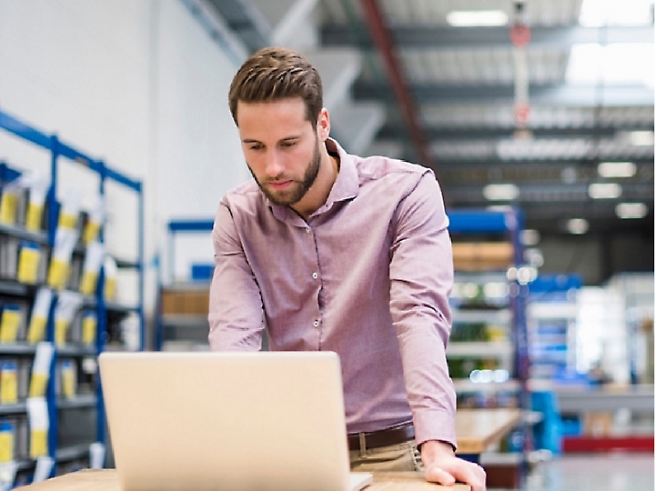 A man in a purple shirt works on a laptop in a warehouse with shelves lined with boxes.