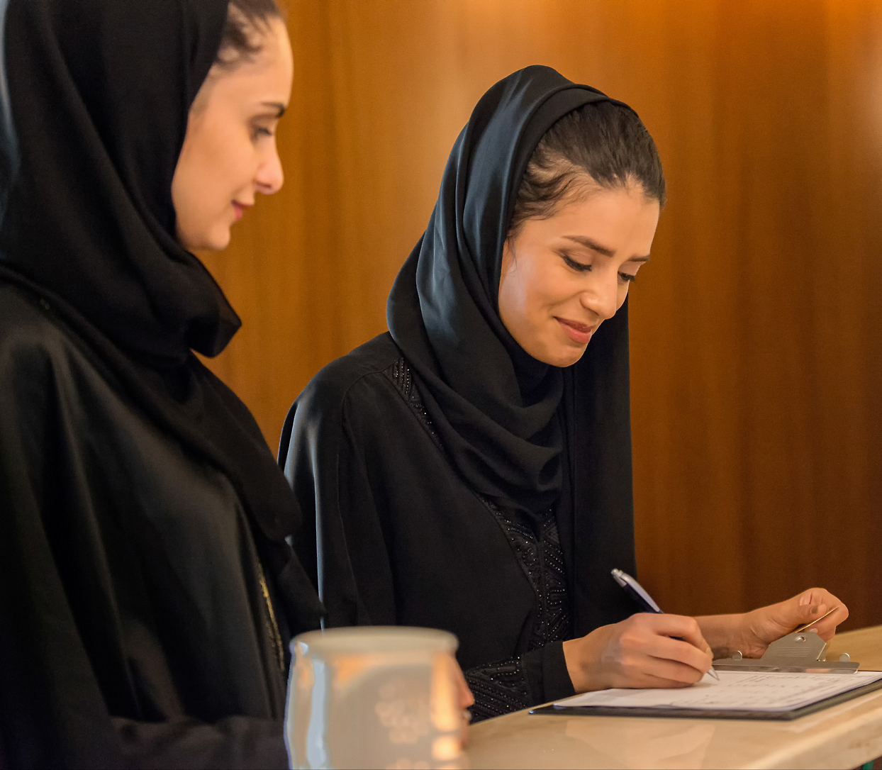 A person in black headscarf writing on a piece of paper another watching her