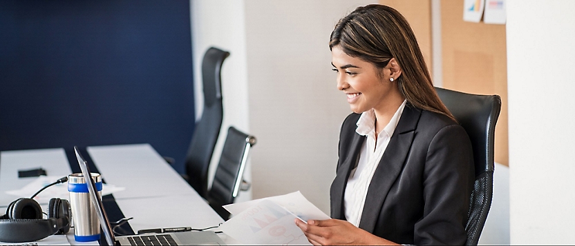 A woman in a business suit is sitting at a desk, smiling and holding papers while working on a laptop in an office environment.