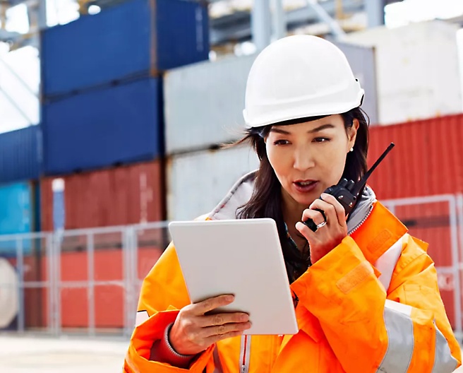 A ground staff wearing safety jacket holding tablet and talking using walkie talkie.