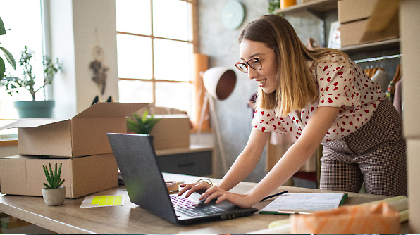 A woman wearing glasses is working on a laptop in an office with boxes and plants around her.