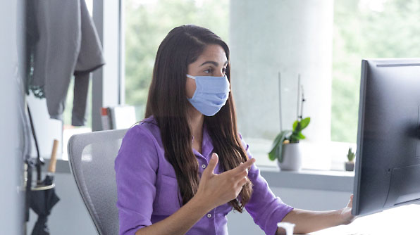 A woman wearing a blue face mask sits at a desk, gesturing with her hand while looking at a computer screen.