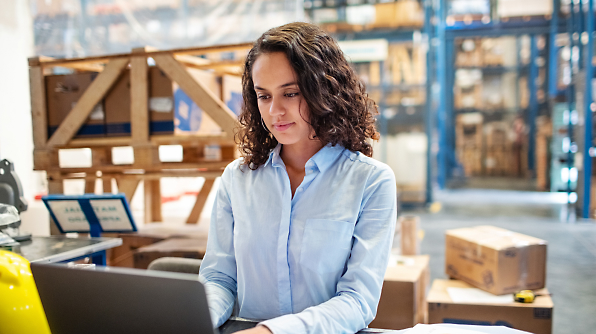 A woman in a blue shirt is working on a laptop in a warehouse filled with boxes and crates.