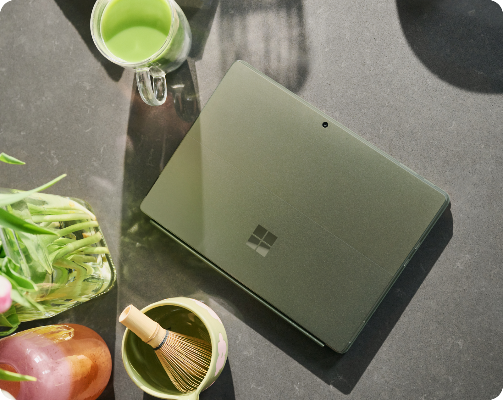 A closed green tablet with a Windows logo lies on a table next to a mug of green liquid