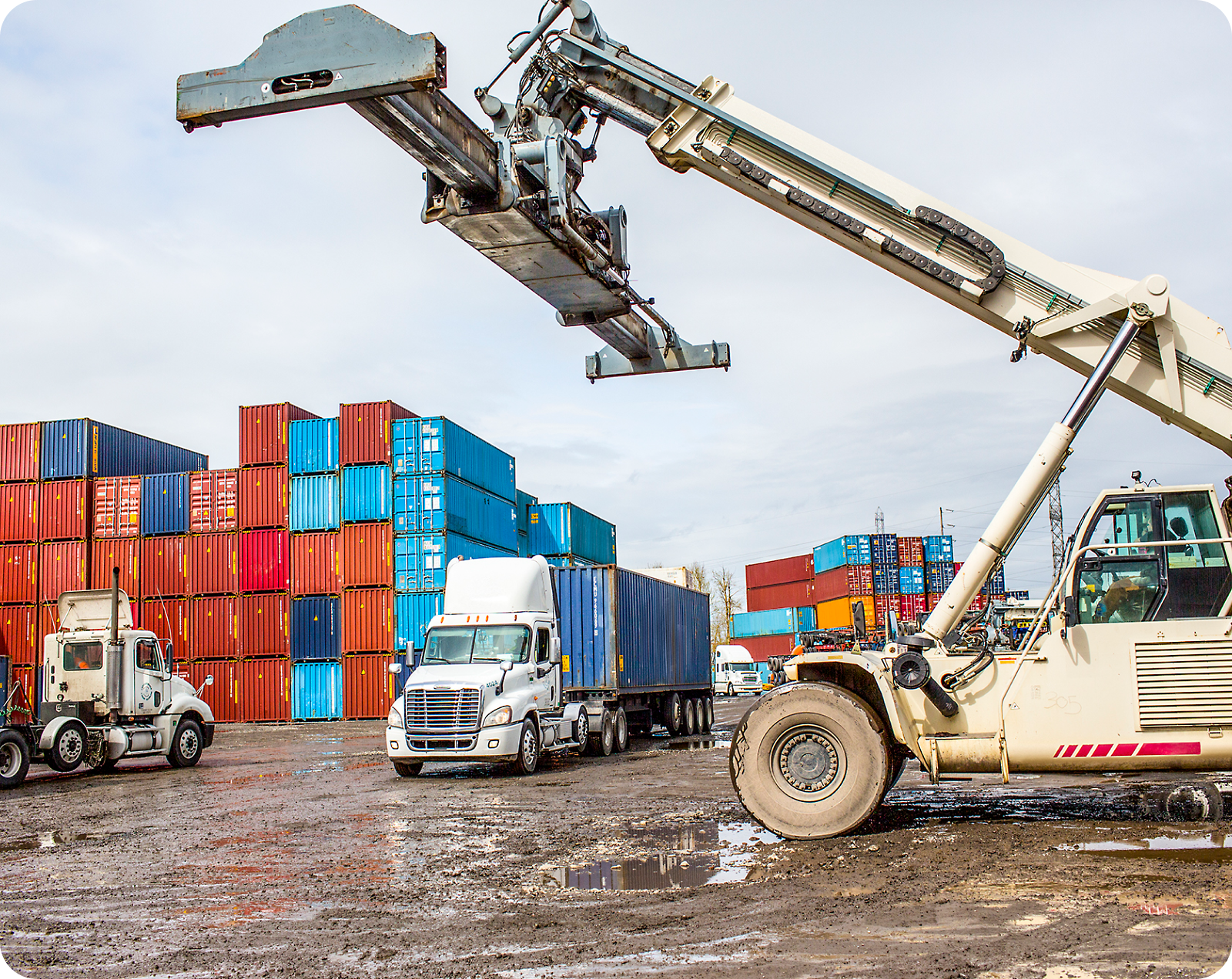 A large machine moves a shipping container in a yard filled with stacked containers and tractor-trailers on a muddy surface.