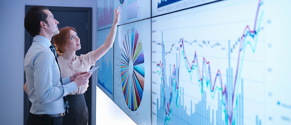 Two person's standing in front of a large screen with a chart