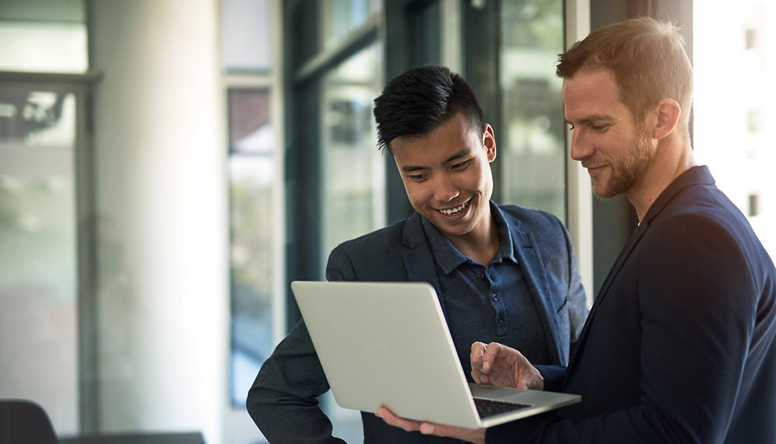 Two men in business attire are standing and looking at a laptop screen together, both smiling, in an office setting.