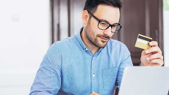 A man wearing glasses is sitting at a desk, holding a credit card in his right hand and looking at a laptop screen