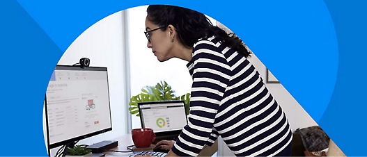 Person wearing a striped shirt, working at a desk with two computer monitors, a red mug, and a plant in the background.