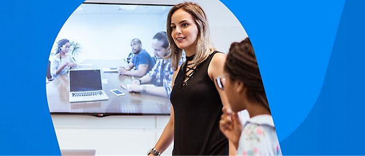 A woman stands in front of a screen displaying a video conference with three people. A young girl stands nearby