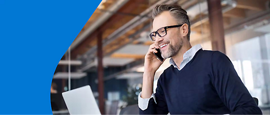 Smiling man with glasses talking on a phone in an open, modern office space with a laptop in front of him.