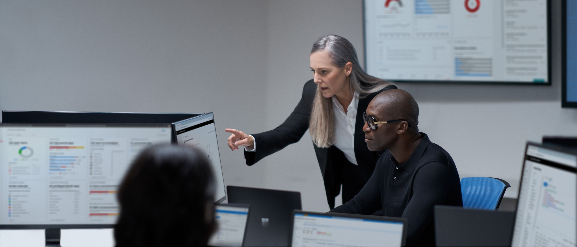 Eine Frau zeigt auf einen Computerbildschirm und steht neben einem sitzenden Mann in einem Büro mit mehreren Monitoren, die Daten anzeigen.