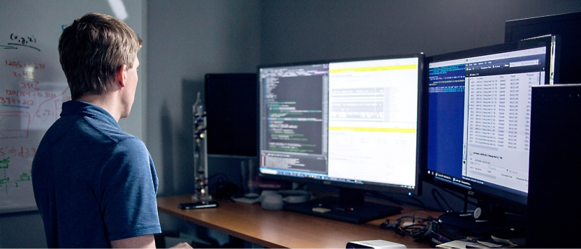 A man in a blue shirt is standing in front of two monitors.