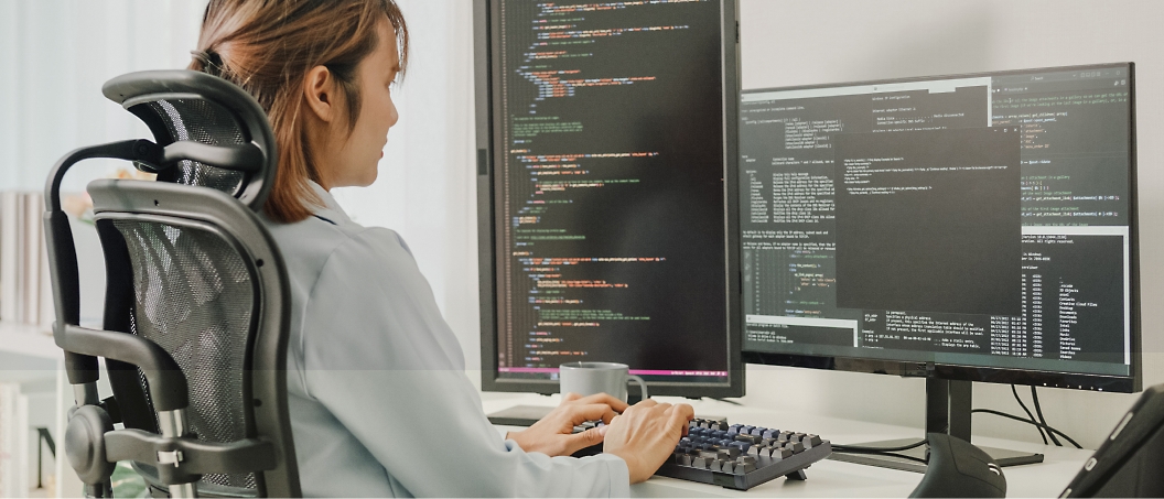 A person sits at a dual-monitor setup in an office chair, typing code on their keyboard