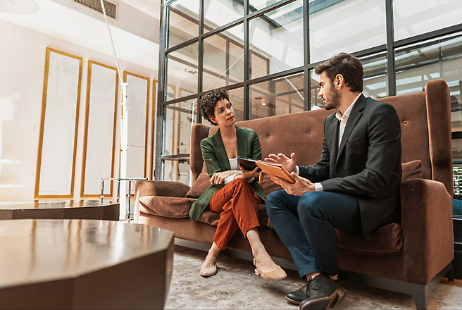 Two professionals sit on a couch in an office, engaged in a discussion while looking at a clipboard.