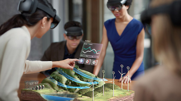 Four people wearing augmented reality headsets interact with 3D model of a landscape that includes wind turbines and solar panels.