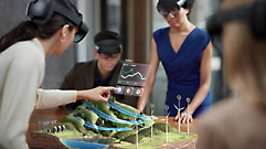 Four people wearing augmented reality headsets interact with 3D model of a landscape that includes wind turbines and solar panels.