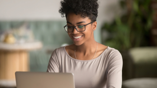 A person with glasses and curly hair is smiling while working on a laptop.