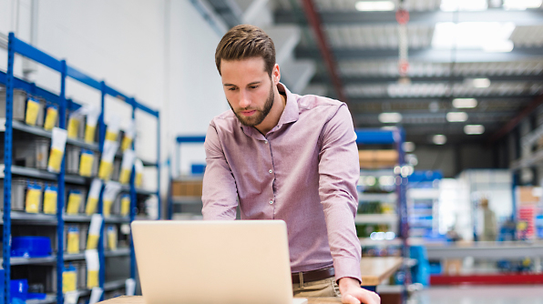 A man in a warehouse setting stands and works on a laptop placed on a desk, surrounded by shelves filled with boxes.