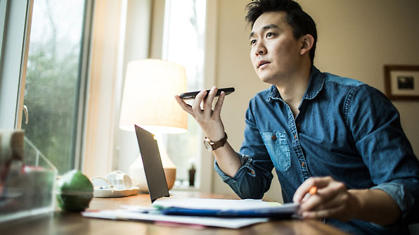 A person is speaking into a phone while working on a laptop at a desk.