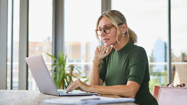 A woman in glasses works on a laptop at a table in a modern, bright room with large windows.