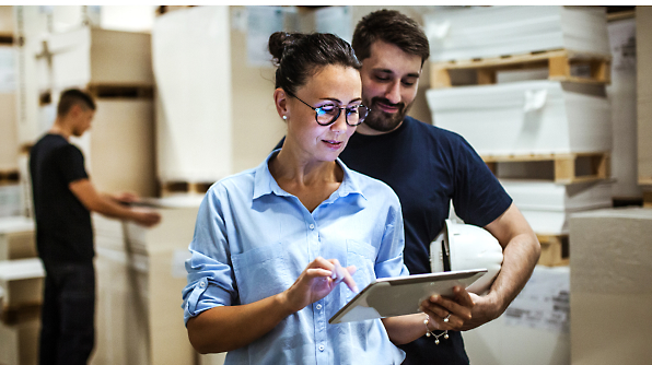 A woman is using a tablet while a man holding a white hard hat stands beside her, looking at the tablet in a warehouse. 