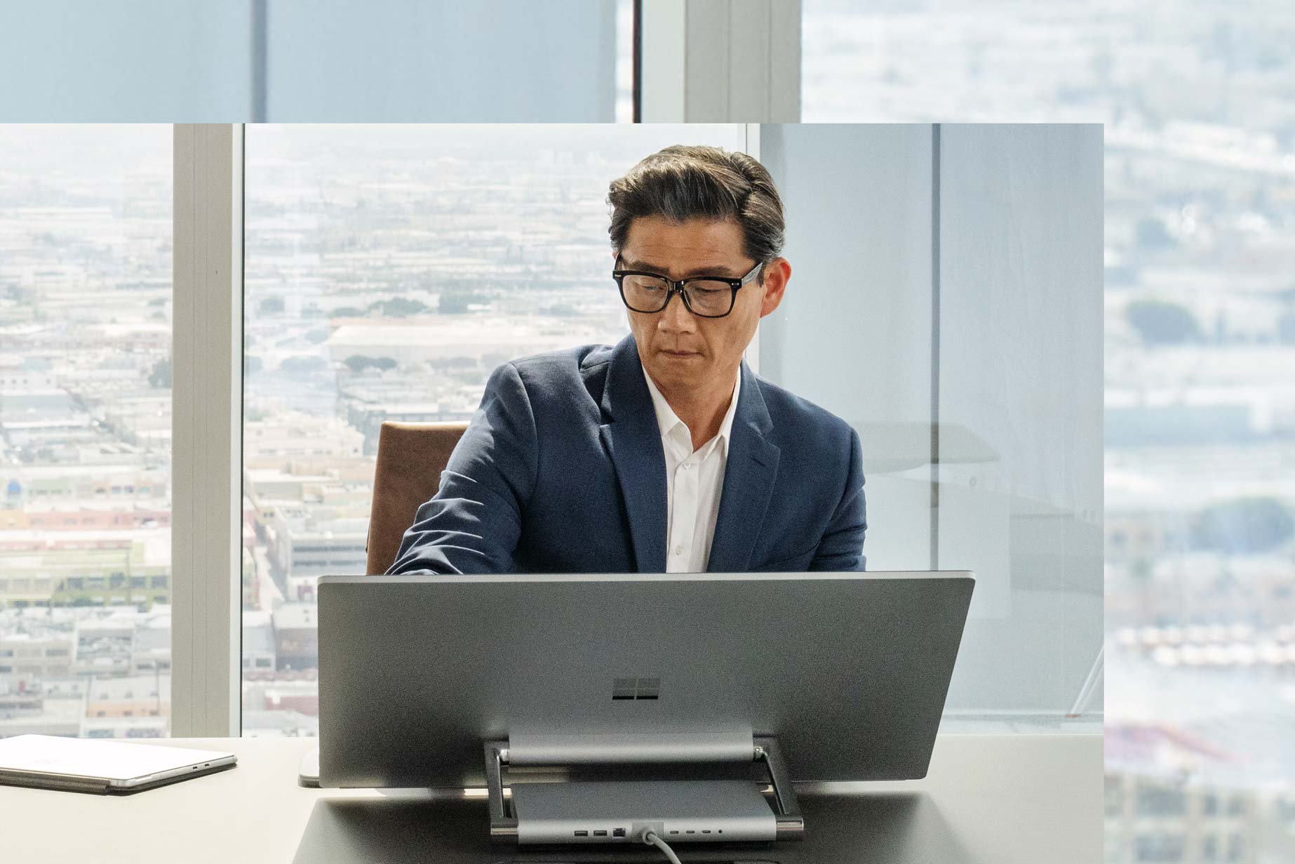 街並みの一部が見える窓の前で Surface Studio 2+ に向かって座る男性。